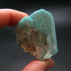 Puede incluir: Un cristal de amazonita verde con una textura rugosa y natural. El cristal tiene una forma puntiaguda y es parcialmente translúcido.