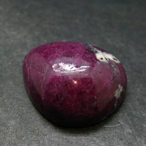 Könnte beinhalten: Ein polierter, abgerundeter Rubin-Edelstein mit einem tiefen Magenta- und Violettton. Der Stein hat eine glatte, reflektierende Oberfläche mit sichtbaren natürlichen Einschlüssen, einschließlich kleiner weißer und schwarzer Mineralablagerungen.
