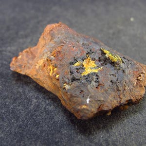 May include: A rough, dark brown rock with yellow mineral deposits. The rock has a rough, uneven surface and is covered in small, yellow crystals.