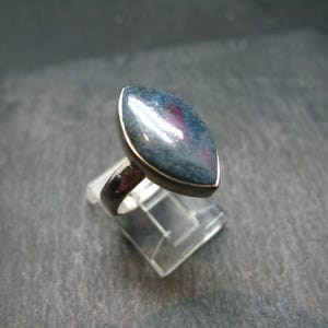 Op de afbeelding: Zilveren ring met een grote, ovaalvormige edelsteen in blauwgroene tinten met rode accenten. De ring is gezet in een gepolijste zilveren band. Gepresenteerd op een transparante standaard, die het ontwerp en de kleuren van de ring benadrukt.