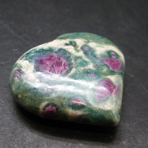 Może przedstawiać: Kamień szlachetny w kształcie serca z mieszanką zielonych, białych i różowo-czerwonych odcieni. Kamień ma wypolerowaną powierzchnię, z różowo-czerwonymi obszarami pojawiającymi się jako wtrącenia w zielonej i białej matrycy. Kamień szlachetny jest prawdopodobnie przedmiotem dekoracyjnym.