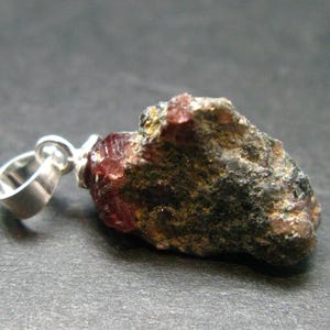 May include: A natural gemstone pendant. The pendant has a rough, dark-coloured stone with red and green hues, attached to a silver-coloured bail. The stone has a textured surface and irregular shape, suggesting it is unpolished.