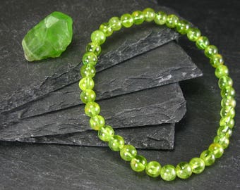 Puede incluir: Un brazalete de cuentas redondas, translúcidas, de color verde lima. Una gema verde en bruto y facetada se encuentra cerca. La joyería se exhibe sobre una superficie oscura y texturizada, creando un contraste que resalta los colores vibrantes.