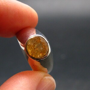 Puede incluir: Un anillo de plata con una piedra preciosa ovalada de color &aacute;mbar. La gema est&aacute; engastada en un bisel, y el anillo se sostiene entre los dedos sobre un fondo oscuro. La banda del anillo es lisa y pulida.