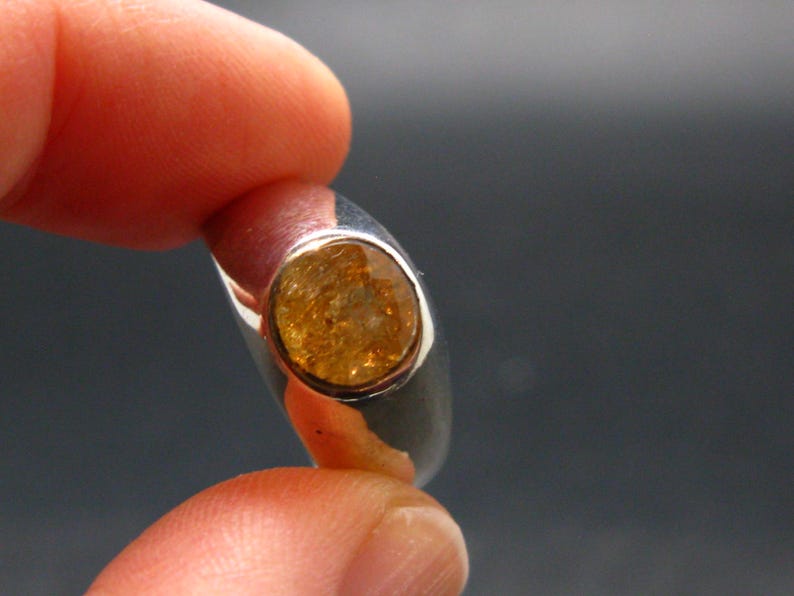 Puede incluir: Un anillo de plata con una piedra preciosa ovalada de color &aacute;mbar. La gema est&aacute; engastada en un bisel, y el anillo se sostiene entre los dedos sobre un fondo oscuro. La banda del anillo es lisa y pulida.