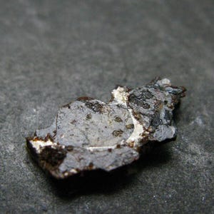 May include: A small, irregular fragment of a meteorite with a dark, textured exterior and a lighter, metallic-looking interior surface. The piece displays a rough, pitted composition with visible mineral inclusions and sharp, jagged edges throughout.