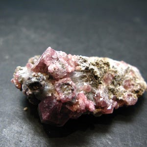 May include: A mineral specimen with a cluster of pink crystals. The crystals have a crystalline structure and are set against a backdrop of a rough, earthy matrix. The color palette includes shades of pink, brown, and white.