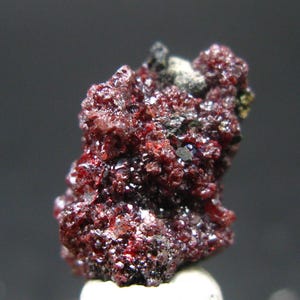 Może przedstawiać: Zbliżenie na głęboko czerwony okaz minerału o szorstkiej, krystalicznej fakturze. Minerał ma ciemną, prawie czarną podstawę z błyszczącymi czerwonymi i białymi refleksami. Okaz jest umieszczony na ciemnoszarym tle.