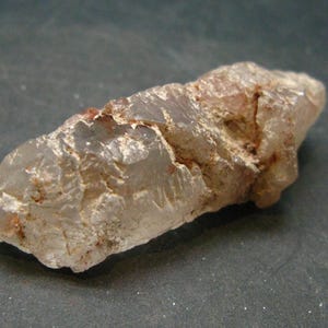 May include: A clear quartz crystal with a rough, natural surface and a reddish-brown hue. The crystal is approximately 5 cm long and has a pointed tip.