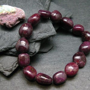 May include: A bracelet composed of polished, deep red, oval-shaped gemstone beads. The beads exhibit a smooth, glossy texture with natural variations in colour and opacity. The bracelet is arranged in a circular shape, showcasing the uniform size and rounded form of each individual stone bead.