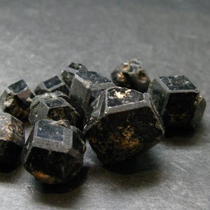 Könnte beinhalten: Eine Ansammlung kleiner, dunkler, undurchsichtiger Mineralkristalle mit geometrischer, facettierter Form. Diese Steine weisen eine tiefschwarze Farbe mit subtilen Texturvariationen und Oberflächenglanz auf und erscheinen als rohe Polyeder.
