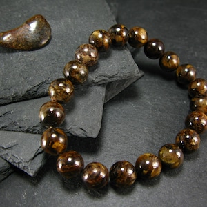 May include: A beaded bracelet featuring large, round, dark brown beads with lighter brown and gold marbling. A matching, polished stone sits to the left of the bracelet. The bracelet is displayed on a dark gray, textured surface.
