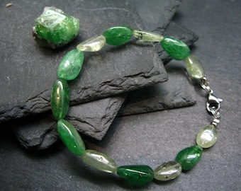 May include: A green and clear gemstone bracelet with a silver clasp. The bracelet is made of oval-shaped beads and is lying on a dark gray surface.