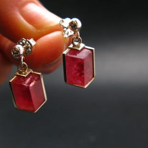 May include: A pair of gold-toned earrings with rectangular, faceted, red gemstones. The earrings have a post and a small decorative element at the top. The gemstones are set in a gold frame.