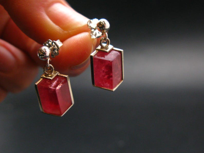 May include: A pair of gold-toned earrings with rectangular, faceted, red gemstones. The earrings have a post and a small decorative element at the top. The gemstones are set in a gold frame.
