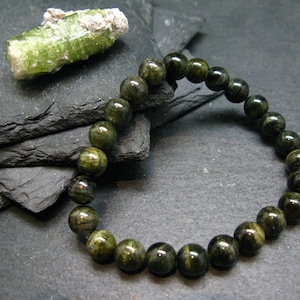 May include: A beaded bracelet featuring dark green, round gemstones. A raw, light green crystal is positioned above the bracelet. The bracelet is displayed on a dark, textured surface. The gemstones are approximately 0.8 cm in diameter.
