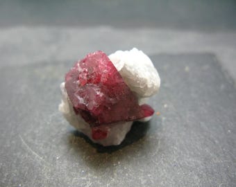 May include: A vibrant, faceted red crystal cluster, possibly a gemstone, is set against a backdrop of white, crystalline material. The red crystal has a rich, saturated color, and the overall composition suggests a natural mineral specimen.