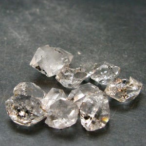 Puede incluir: Una colección de pequeños cristales de cuarzo en bruto, transparentes, con facetas naturales y formas irregulares. Cada cristal muestra una apariencia translúcida con pequeñas inclusiones minerales oscuras visibles.