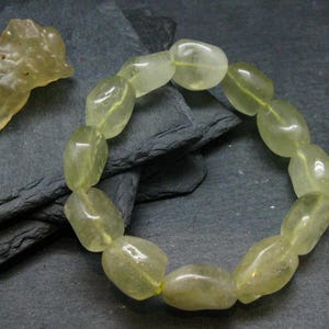 Könnte beinhalten: Ein Armband aus glatten, durchscheinenden, blass gelb-grünen Trommelsteinperlen, begleitet von einem einzelnen, unregelmäßigen, strukturierten Stück rohem, blass gelb-grünem Stein mit einer genarbten Oberfläche.