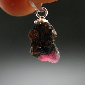Könnte beinhalten: Ein silberner Anhänger mit einem rauen, dunkelbraunen und rosafarbenen Edelstein. Der Edelstein ist ein roher, natürlicher Kristall mit einer unregelmäßigen Form.