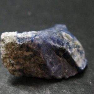 May include: A raw, unpolished mineral specimen featuring a combination of deep blue crystalline sections and rough, textured greyish-white matrix rock. The stone displays irregular edges and a natural, jagged surface with varying levels of opacity.
