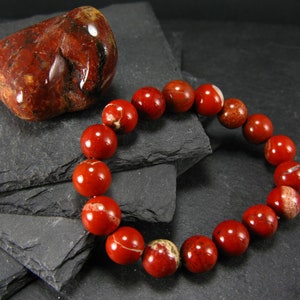 Puede incluir: Un brazalete de cuentas de jaspe rojo con una piedra de jaspe rojo a juego. El brazalete está hecho de cuentas redondas y está sobre un fondo gris oscuro.