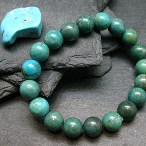 Peut inclure: Un bracelet élastique circulaire composé de perles de turquoise rondes et polies dans des tons variés de bleu sarcelle, de vert et de bleu avec des veines sombres, accompagné d'une pièce de pierre turquoise lisse en forme de croissant.