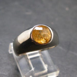 Puede incluir: Un anillo de plata con una gema grande y redonda de color &aacute;mbar. El anillo tiene una banda ancha y un acabado pulido, que muestra los tonos c&aacute;lidos de la gema. La gema est&aacute; engastada en un engaste de bisel.