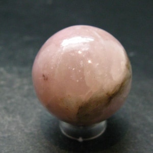 Puede incluir: Una piedra esférica pulida con una superficie lisa y brillante. La esfera presenta un color rosa pálido suave con áreas translúcidas e inclusiones minerales distintivas de color marrón oscuro a negro que crean un patrón natural veteado.