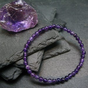 Peut inclure: Un bracelet en améthyste violette avec de petites perles rondes. Le bracelet est sur un fond gris foncé.