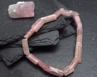 May include: A strand of pink tourmaline beads, each bead is a rectangular prism with striations. The beads are arranged in a loose circle on a dark gray surface.