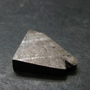 May include: A triangular fragment of a metallic meteorite with a dark, weathered exterior. The surface displays a visible Widmanstätten pattern of intersecting lines and crystalline structures, with a small notch on one of the edges.