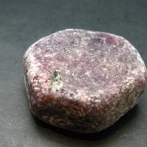 May include: A rounded, natural stone with a mottled appearance. The stone's color is primarily a deep reddish-purple, with lighter, almost white, patches. The surface has a rough, textured feel, and the stone appears to be solid and heavy.