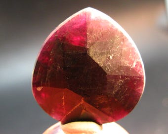May include: A heart-shaped faceted gemstone in a deep, translucent red hue. The stone features multiple geometric facets that catch the light, displaying variations in color intensity from dark crimson to lighter magenta tones throughout.