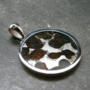 Puede incluir: Un colgante circular con un marco de metal plateado pulido y una reasa. La incrustación central consiste en fragmentos de piedra de color marrón oscuro, translúcidos y opacos, incrustados en una matriz metálica reflectante de color plata.