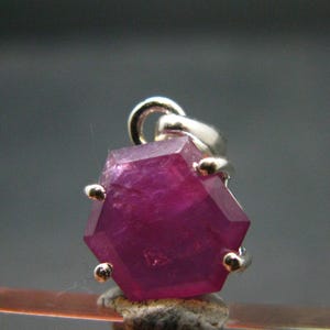 May include: A hexagonal, deep pink gemstone pendant set in a silver-toned metal prong setting. The stone features a flat, faceted surface with visible internal inclusions and a polished finish, suspended from a small, circular silver-toned metal bail.