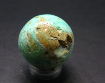 Puede incluir: Una esfera de piedra de color turquesa con parches marrones y tostados. El orbe tiene una superficie rugosa y texturizada y se asienta sobre una base plateada. El color de la piedra es una mezcla de verde y azul.