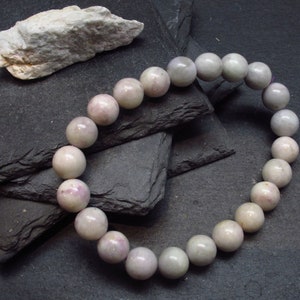 May include: A light pink beaded bracelet with large, round beads. The bracelet is made of polished stones and is displayed on a dark gray surface. A piece of light-colored stone is in the background.