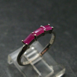 May include: A silver-toned ring featuring three rectangular, baguette-cut vibrant pinkish-red gemstones set in a row. The stones are held in place by small metal prongs, and the band is a smooth, polished metal that curves to form a circular shape.