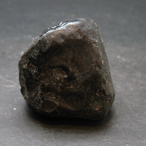 May include: A dark, irregularly shaped stone with a rough, textured surface. The stone is a deep black colour, with subtle variations in tone and highlights. The overall shape is rounded, with a slightly glossy appearance in certain areas.