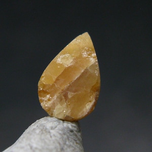 May include: A teardrop-shaped, golden-yellow gemstone with a textured surface. The gem is set against a dark gray background, showcasing its natural color variations and internal patterns. The stone is resting on a gray rock.