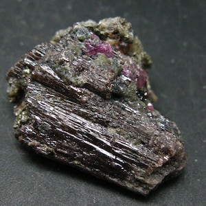Puede incluir: Un espécimen mineral de color marrón oscuro y negro con un grupo de pequeños cristales rosados. El mineral tiene una textura estriada.