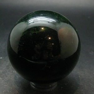 Puede incluir: Una piedra esférica pulida de color verde oscuro profundo. La superficie es lisa y reflectante, mostrando reflejos de luz y sutiles variaciones internas dentro del material denso y opaco. El objeto tiene una forma perfectamente redonda.