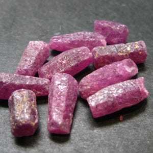 May include: A collection of small, raw, cylindrical ruby crystals with a vibrant magenta-pink hue. Each piece displays a rough, textured surface with natural crystalline facets and subtle variations in colour intensity across the group.