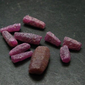 May include: A collection of eleven raw, unpolished ruby crystals in varying shades of deep pink and magenta. The stones feature irregular, elongated, and cylindrical shapes with textured, matte surfaces, scattered across the dark, flat surface.