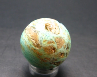 Puede incluir: Una gema esférica pulida con aspecto moteado. La piedra presenta una mezcla de tonos turquesa y marrón, con una superficie texturizada. La gema se exhibe sobre un soporte transparente, mostrando su color y patrón únicos.