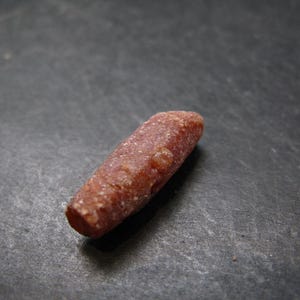 May include: A close-up of a rough, reddish-brown gemstone with a cylindrical shape. The stone has a textured surface and appears to be a natural mineral. The background is a dark, textured surface.