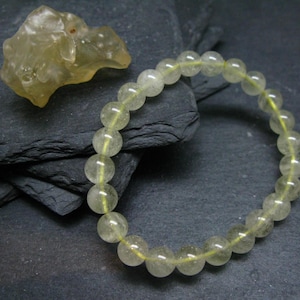 Puede incluir: Una pulsera de cuentas amarillas pálidas y translúcidas. También hay una piedra a juego de forma irregular. La pulsera está hecha de cuentas redondas ensartadas en un cordón amarillo. El fondo es una superficie oscura y texturizada.