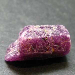 Puede incluir: Primer plano de una gema de rubí en bruto sin cortar. El rubí es de un color rojo-rosado intenso y vibrante con una superficie texturizada. Pequeñas motas doradas y verdes son visibles en la superficie, lo que aumenta su apariencia natural.