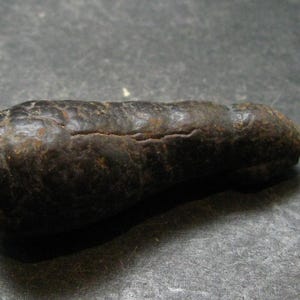 Puede incluir: Un objeto de color marrón oscuro, alargado y redondeado con una superficie rugosa y texturizada. El artículo presenta una fisura o grieta horizontal distintiva a lo largo de su longitud, con variaciones de color sutiles y un aspecto desgastado.